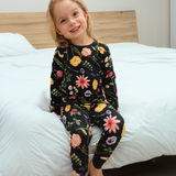Et barn sidder på en seng iført et langærmet sort pyjamassæt med et farverigt blomstermønster.